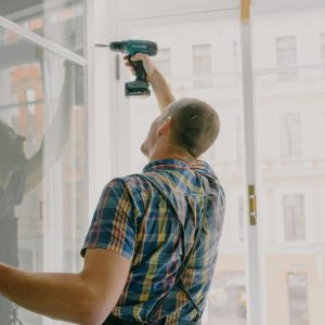 Back view of worker in checkered shirt drilling window frame during renovation process in apartment